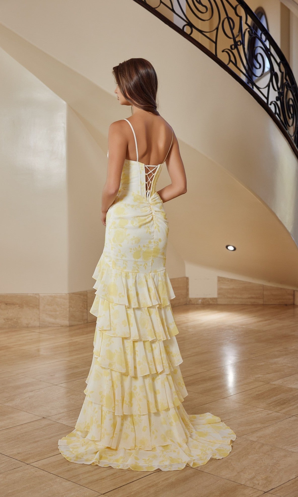 A woman in the Nox Anabel T1863 long prom dress, featuring a light yellow tiered ruffle skirt, stands with her back to the camera on a polished floor near an ornate curved staircase.